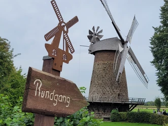 Historische Windmühle aus Stein mit Holzflügeln, umgeben von grüner Landschaft und Hinweisschild.