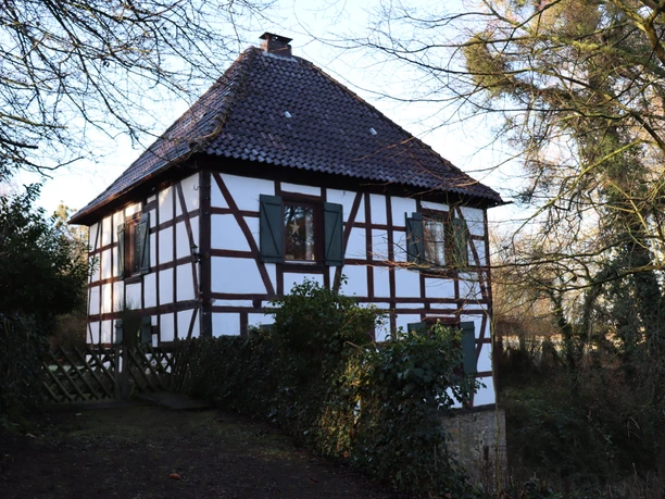 Fachwerkgebäude mit roten Dachziegeln, umgeben von Bäumen; ruhige ländliche Atmosphäre.