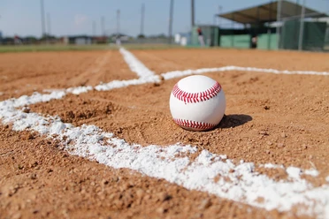 Baseballfeld mit Mittelkreis und Basenlinien, im Vordergrund liegt ein weißer Ball auf sandigem Boden.
