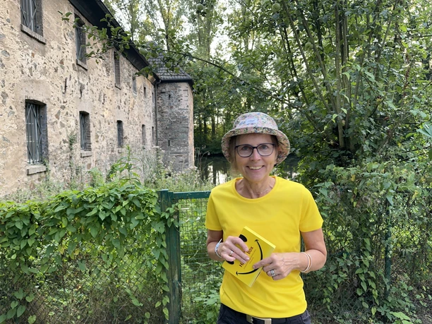 Lachwanderung mit Claudia Thienenkamp, Langenfeld Eine lächelnde Person im gelben T-Shirt und buntem Hut steht vor einem historischen Gebäude.