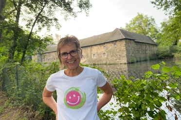 Lachwanderung mit Claudia Thienenkamp, Langenfeld Claudia Thienenkamp, lächelnd im Freien, vor einem steinernen Gebäude in Langenfeld, am Wasser.