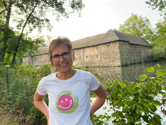 Lachwanderung mit Claudia Thienenkamp, Langenfeld Claudia Thienenkamp, lächelnd im Freien, vor einem steinernen Gebäude in Langenfeld, am Wasser.