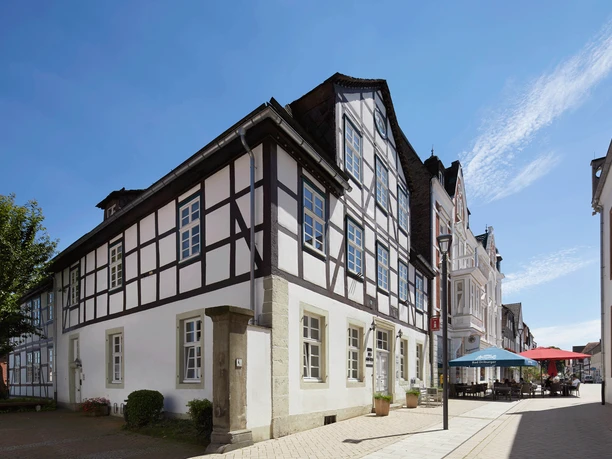 Haus_des-Gastes_2019_Blickrichtung-Marktplatz.png Fachwerkhaus am Marktplatz vor blauem Himmel; nebenan ein Café mit bunten Sonnenschirmen.