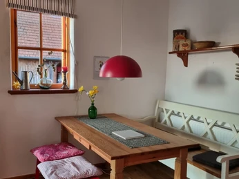 Ein Esstisch mit Holzbank und rotem Hocker, geschmückt mit gelber Vasenblume unter roter Hängelampe.