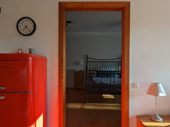 Roter Kühlschrank, Holztürrahmen spiegelnd ein Schlafzimmer mit Eisenbett, Holzboden, Wanduhr.