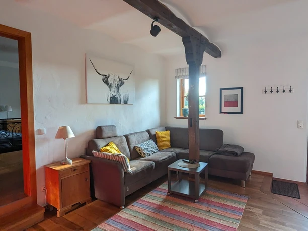 Gezellige woonkamer met bank, houten balken en kunstwerk van een koe. Deur leidt naar het zonnige terras.