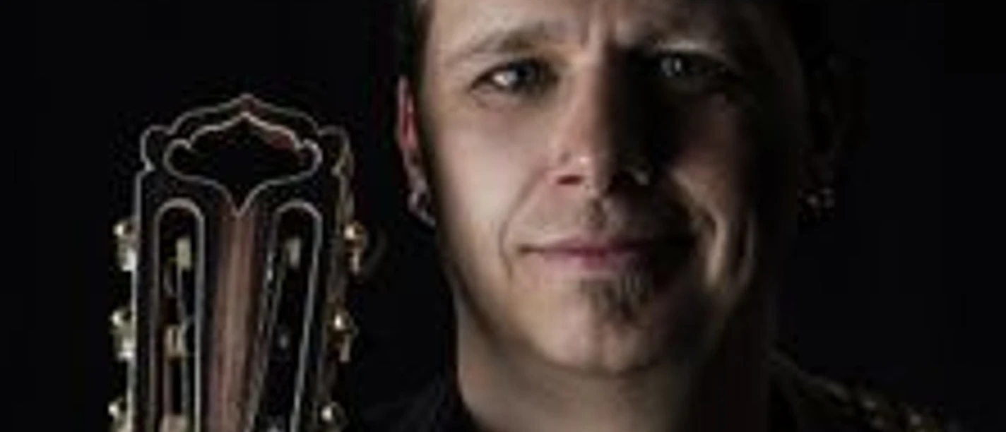 Ein Musiker mit geflochtenem Haar hält eine akustische Gitarre an der Schulter, sanft beleuchtet.