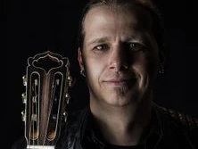 Ein Musiker mit geflochtenem Haar hält eine akustische Gitarre an der Schulter, sanft beleuchtet.