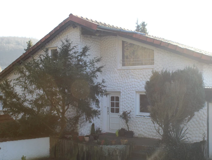 Außenansicht Gästehaus SL Immobilien Zweistöckiges Gebäude mit weißer, strukturiert wirkender Fassade, umgeben von Bäumen und Pflanzen.