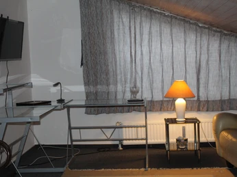 Gästehaus SL Immobilien Dachzimmer mit Glastisch und Stehlampe neben einem Sofa; Fernseher an der Wand, Fenster mit Vorhängen.