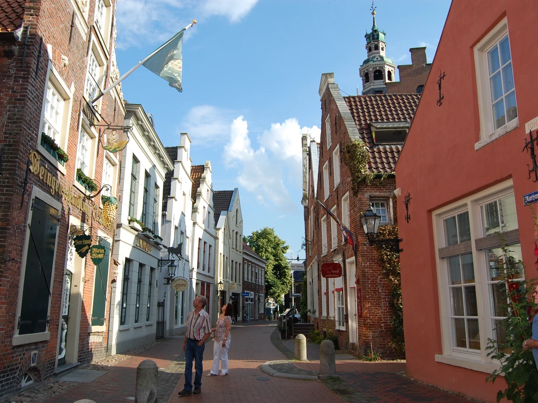 2106_altstadt_von_leer-original Gepflasterte Gasse in der Altstadt von Leer mit historischen Backsteinhäusern und Passanten bei Sonne.