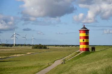 Pilsumer Leuchtturm Roter und gelber Leuchtturm auf grünem Deich mit weitem Himmel und Windrädern im Hintergrund.
