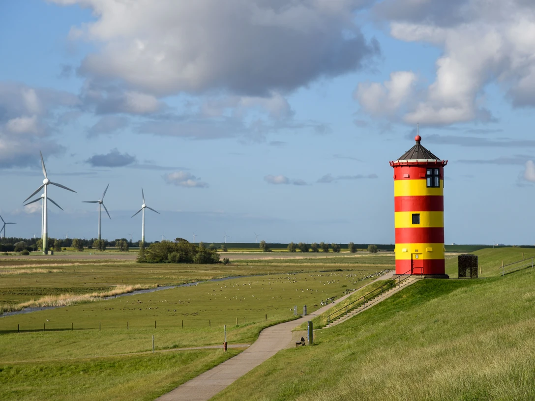 Pilsumer Leuchtturm Roter und gelber Leuchtturm auf grünem Deich mit weitem Himmel und Windrädern im Hintergrund.