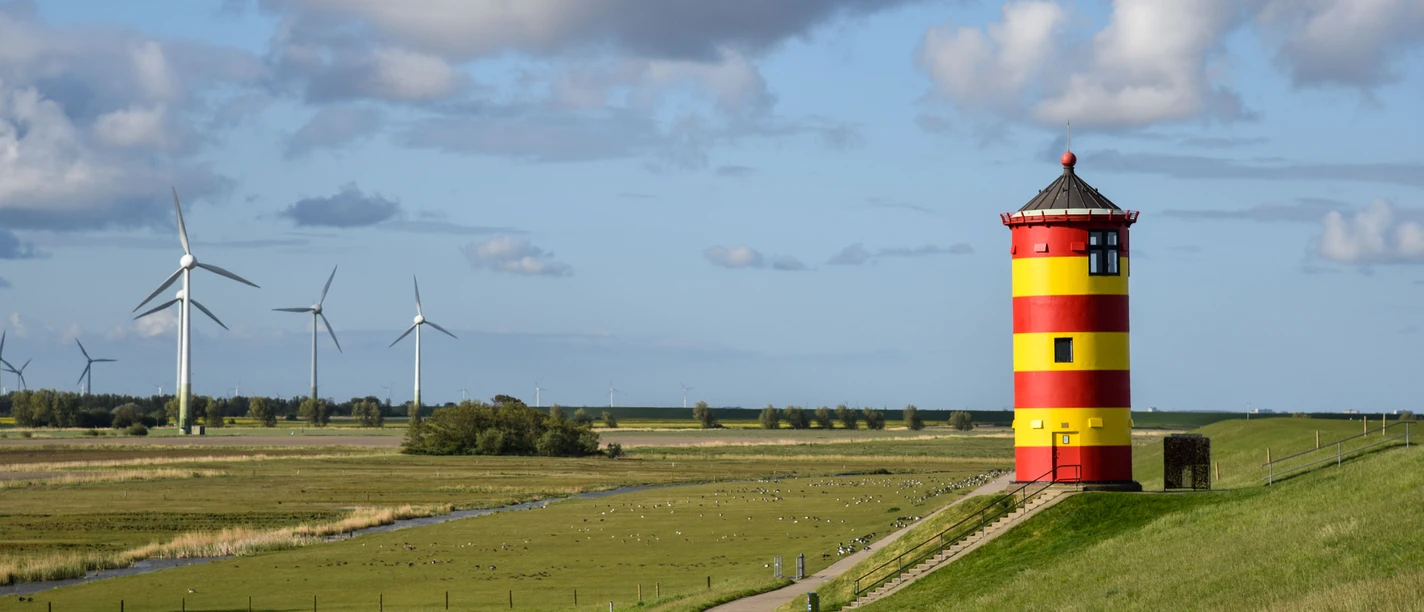 Pilsumer Leuchtturm Roter und gelber Leuchtturm auf grünem Deich mit weitem Himmel und Windrädern im Hintergrund.