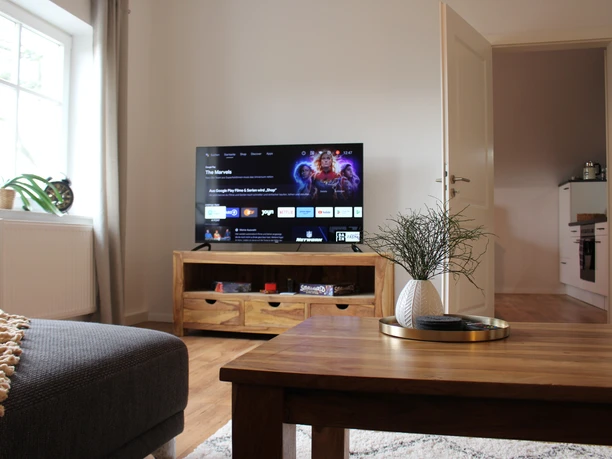 IMG_1112.JPG Ein moderner Wohnraum mit grauem Sofa, Holztisch und Flachbildfernseher vor beige gestrichenen Wänden.