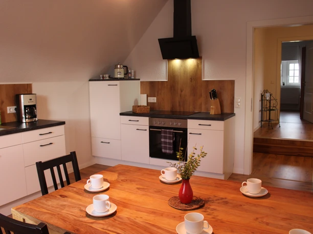 Modern fitted kitchen with wooden table, four white cups, chairs, stove and open door view.