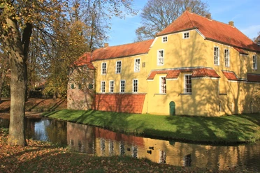 Manningaburg Pewsum Gelbes Backsteingebäude mit rotem Ziegeldach, umgeben von Wassergraben und herbstlichen Bäumen.