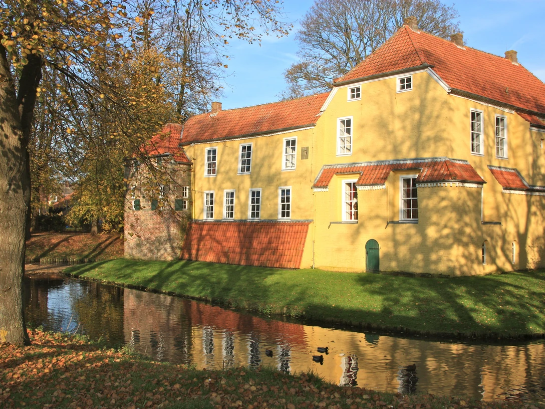 Manningaburg Pewsum Gelbes Backsteingebäude mit rotem Ziegeldach, umgeben von Wassergraben und herbstlichen Bäumen.