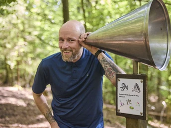 LeistruperWald-LeistruperWaldRoute-LeistruperWald-LTMGmbH-HirschmeierMediaGmbH&CoKG_024.jpg Mann lauscht im Leistruper Wald einem Waldhörrohr. Umgeben von grünen Bäumen und Tageslicht.