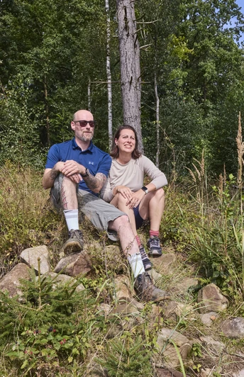 Zwei Personen sitzen auf einer Steinbank im Wald und genießen die sonnige Atmosphäre umgeben von Bäumen.