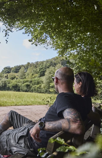 Dörentrup-NABULehrpfad-Teutoburger Wald-LTMGmbH-HirschmeierMediaGmbH&CoKG_025.jpg Ein Paar sitzt auf einer Bank am Waldrand und blickt über eine weitläufige Wiesenlandschaft.