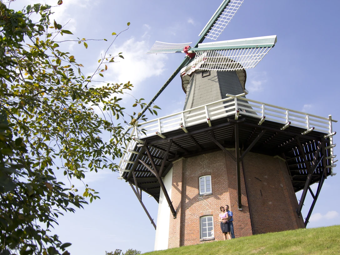 Mühle Greetsiel Backsteinerne Windmühle mit weißen Flügeln vor blauem Himmel, zwei Personen stehen am Fuß des Bauwerks.