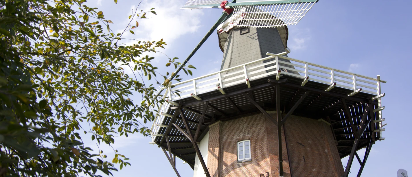 Mühle Greetsiel Backsteinerne Windmühle mit weißen Flügeln vor blauem Himmel, zwei Personen stehen am Fuß des Bauwerks.