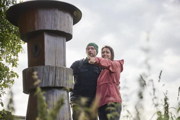 Kalletal-TallerBergrunde-TeutoburgerWald-LTMGmbH-HirschmeierMediaGmbH&CoKG_013.jpg Ein Paar steht lächelnd vor einem großen Holzpfosten im Freien und zeigt in die Ferne.