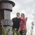 Kalletal-TallerBergrunde-TeutoburgerWald-LTMGmbH-HirschmeierMediaGmbH&CoKG_013.jpg Ein Paar steht lächelnd vor einem großen Holzpfosten im Freien und zeigt in die Ferne.