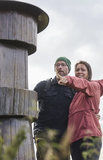 Kalletal-TallerBergrunde-TeutoburgerWald-LTMGmbH-HirschmeierMediaGmbH&CoKG_013.jpg Ein Paar steht lächelnd vor einem großen Holzpfosten im Freien und zeigt in die Ferne.