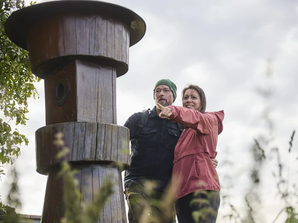 Kalletal-TallerBergrunde-TeutoburgerWald-LTMGmbH-HirschmeierMediaGmbH&CoKG_013.jpg Ein Paar steht lächelnd vor einem großen Holzpfosten im Freien und zeigt in die Ferne.
