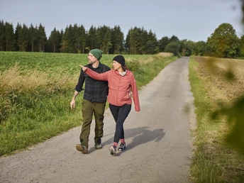 Kalletal-TallerBergrunde-TeutoburgerWald-LTMGmbH-HirschmeierMediaGmbH&CoKG_027.jpg Ein Paar spaziert auf einem ländlichen Weg, flankiert von Feldern und Wäldern im Teutoburger Wald.