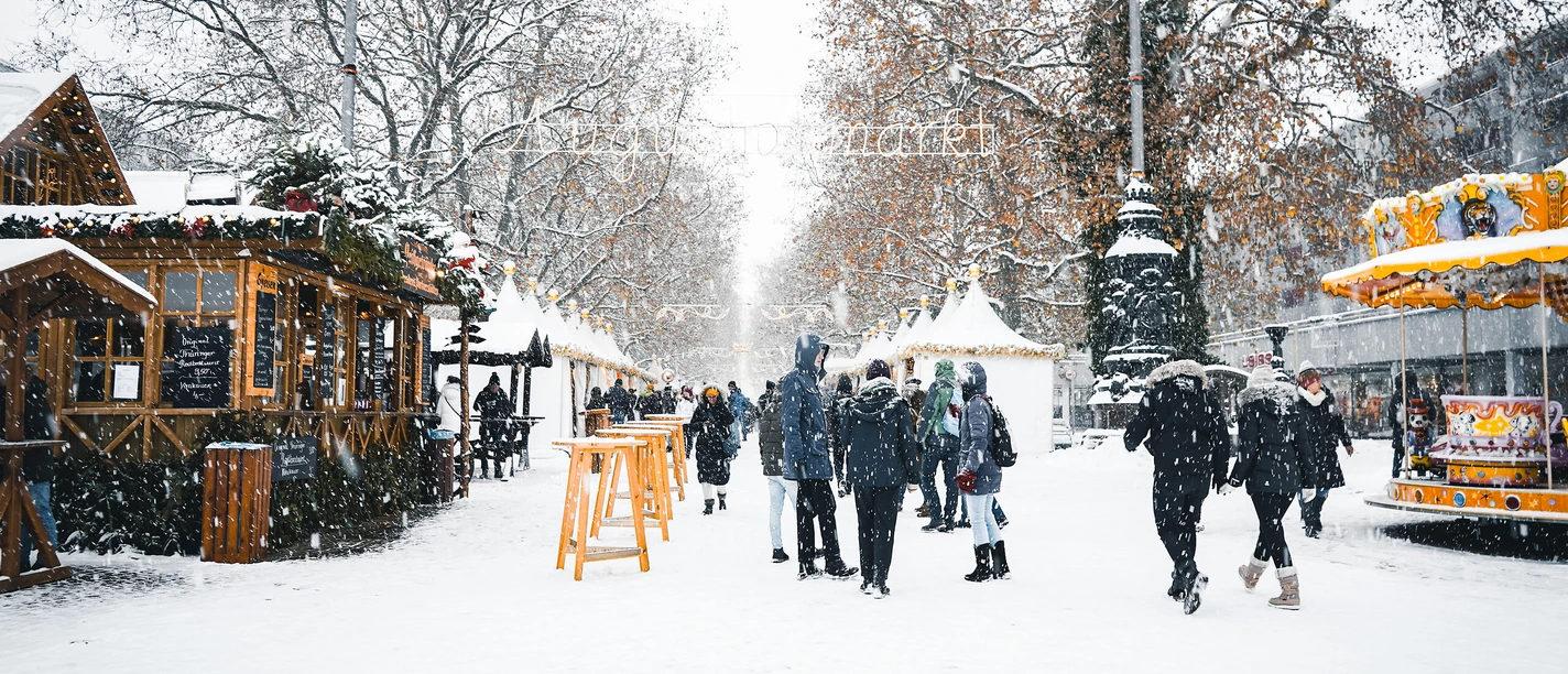 Augustusmarkt auf der Hauptstraße in Dresden.jpg Winterlicher Markt mit Ständen und Menschengruppen im Schnee unter kahlen Bäumen.