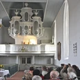 Kirche in Groothusen Gäste sitzen in Kirchenbänken und blicken auf die große weiße Orgel der Kirche in Groothusen.