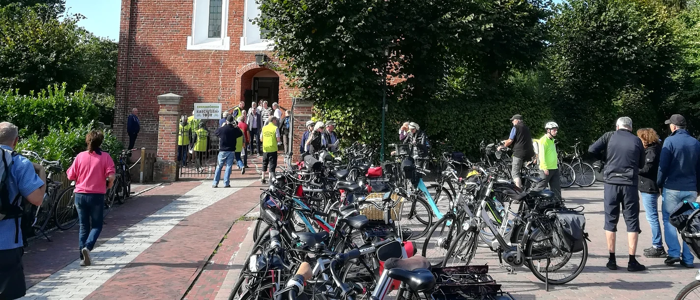 Krummhörner Kirchturm Tour 2018 Radfahrerinnen und Radfahrer versammeln sich vor der Backsteinkirche Jennelt bei sonnigem Wetter.