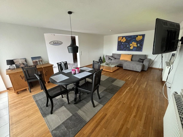 Wohnzimmer.jpg Geräumiges Wohnzimmer mit Holzboden, Esstisch, grauem Sofa und großer Wandmalerei in warmen Farben.