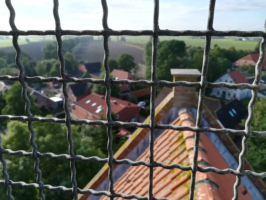 Krummhörner Kirchturm Tour: Blick auf Eilsum Blick durch das Schutzgitter eines Kirchturms auf rote Ziegeldächer und grüne Landschaft in Eilsum.