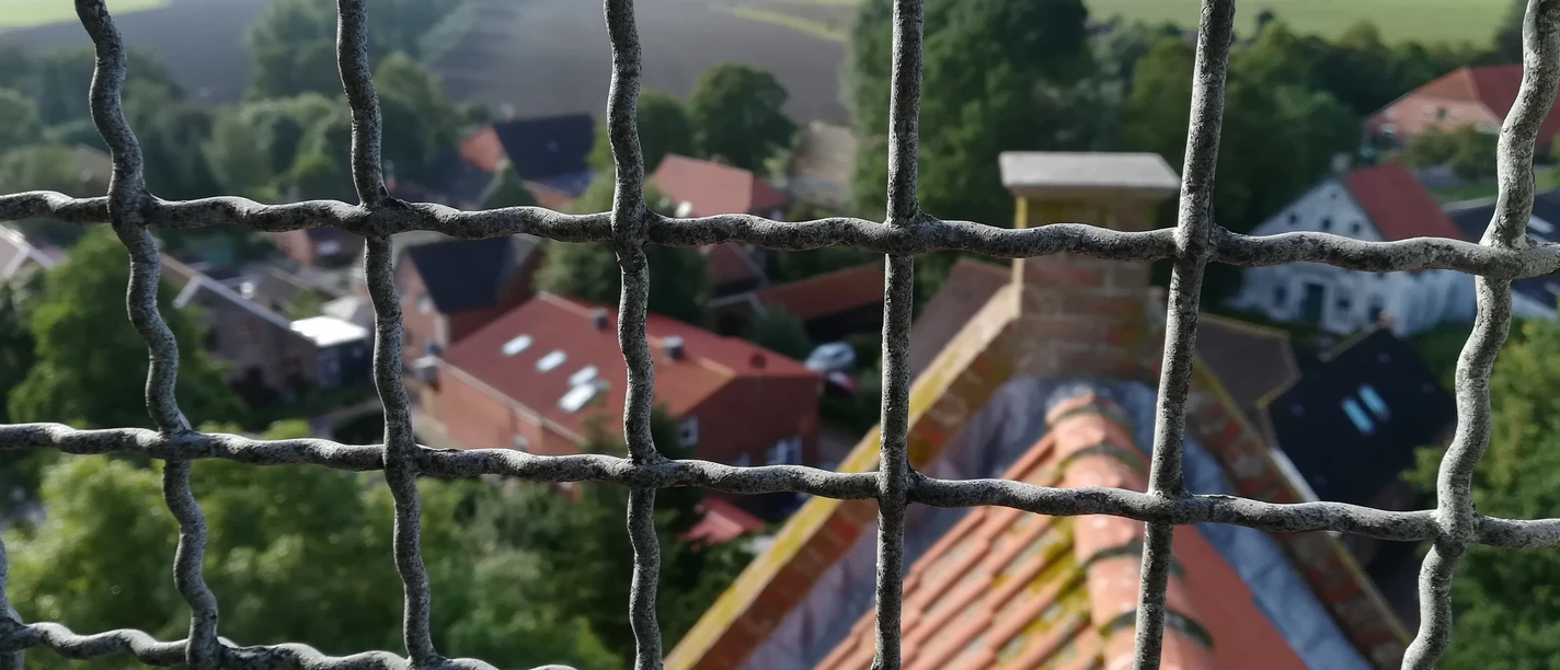 Krummhörner Kirchturm Tour: Blick auf Eilsum Blick durch das Schutzgitter eines Kirchturms auf rote Ziegeldächer und grüne Landschaft in Eilsum.
