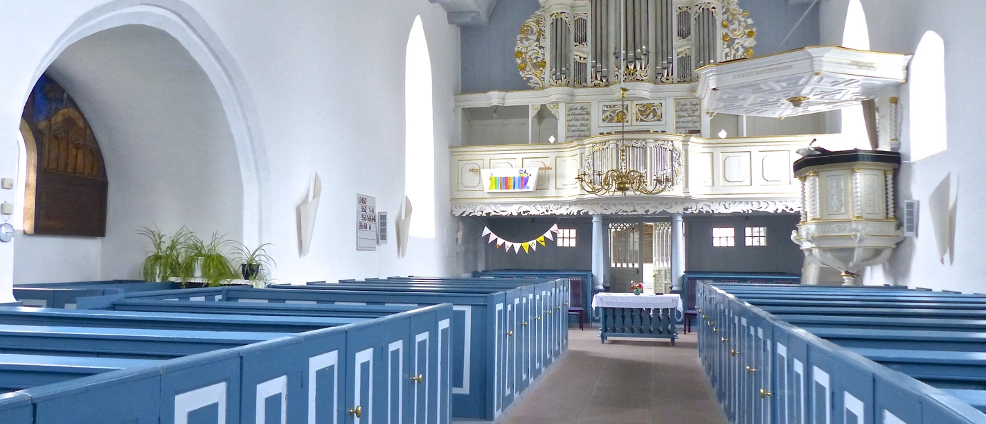 Kirche Manslagt Heller Kircheninnenraum mit blauen Holzbänken, weißer Kanzel und prunkvoller Orgel.
