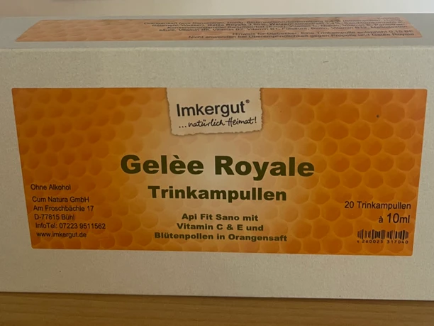 Weiße Verpackung mit orangefarbenem Etikett für Gelee Royale Trinkampullen auf Holzoberfläche.