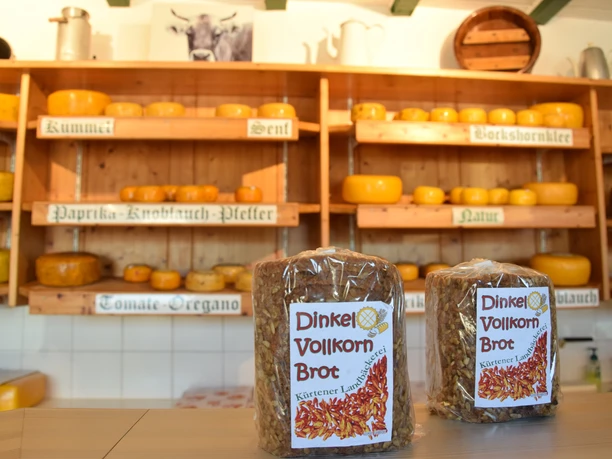 Thomashof Burscheid Zwei Dinkelvollkornbrote vor einem Regal voller Käselaibe in einem rustikalen Geschäft.