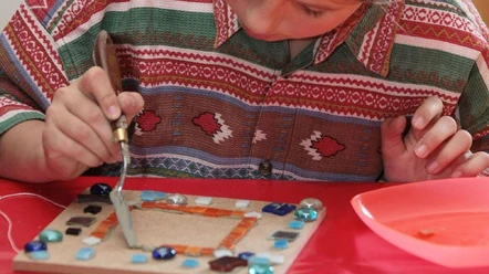 Überraschungswerkstatt 2.jpg Ein Kind verziert konzentriert eine Holzplatte mit bunten Glasstücken und hebt sie sorgsam an.