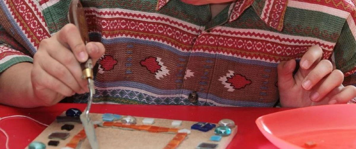 Überraschungswerkstatt 2.jpg Ein Kind verziert konzentriert eine Holzplatte mit bunten Glasstücken und hebt sie sorgsam an.