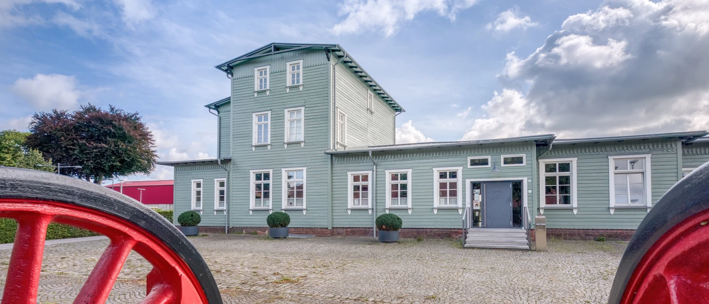 Bahnhof Rahden Historischer Bahnhof Rahden, grünes Gebäude, Dreigeschossturm, Kopfsteinpflaster, blühende Sträucher.