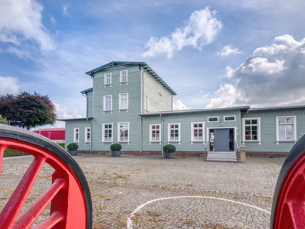 Bahnhof Rahden Historischer Bahnhof Rahden, grünes Gebäude, Dreigeschossturm, Kopfsteinpflaster, blühende Sträucher.