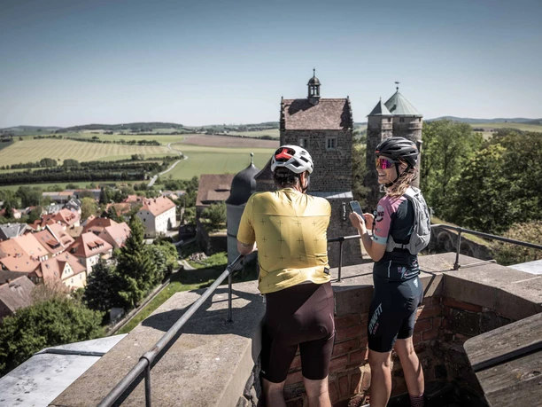 Ausblick von der Burg Stolpen Zwei Radfahrer mit Helmen stehen auf einer Mauer der Burg Stolpen und blicken auf eine malerische Landschaft mit Feldern und einem Dorf mit roten Dächern.