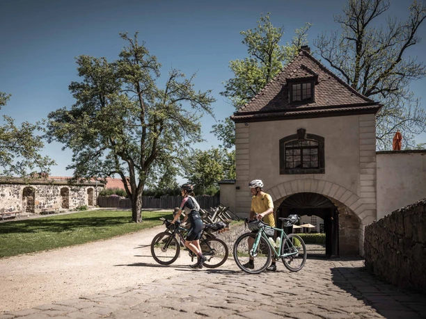 Castle courtyard at Stolpen Castle Two cyclists wearing helmets ride along a cobbled path at Stolpen Castle, surrounded by trees and sunny weather.