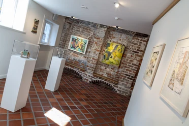 Galerie im Glockenhof Heller Galerieraum mit Backsteinwand, Bodenfliesen, Gemälden und Skulpturen auf Podesten.