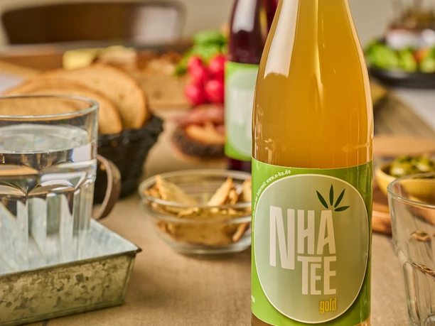 Königs NHA Hanftee Eine goldene Flasche Hanftee mit grünem Etikett steht auf einem gedeckten Tisch mit Snacks.