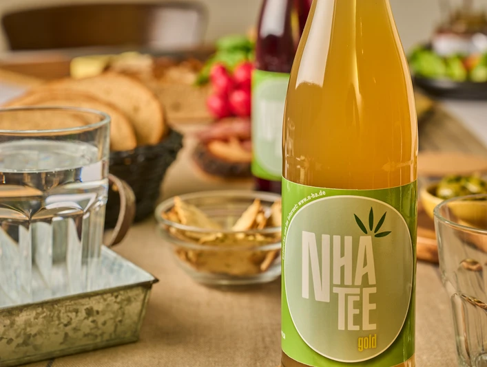 Königs NHA Hanftee Eine goldene Flasche Hanftee mit grünem Etikett steht auf einem gedeckten Tisch mit Snacks.